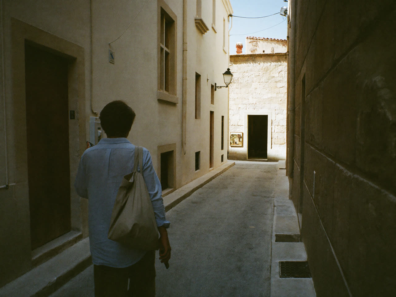 Traveller in a Malta limestone lane searching on their phone for a place to stay, pausing to look toward a lit guesthouse doorway